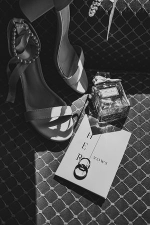 Black and white photo of wedding accessories including high heel shoes, perfume bottle and vows book on textured surface.