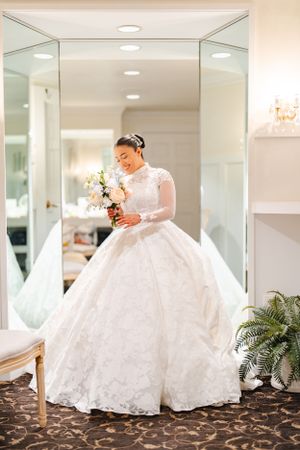 A bride in an elegant white lace ballgown with long sleeves looks at her bouquet in a mirrored bridal suite.
