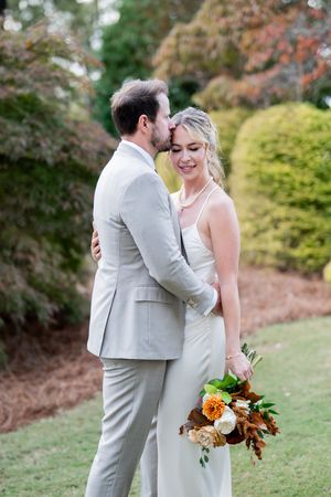 A couple shares an intimate moment outdoors surrounded by autumn foliage and holding a fall-colored bouquet.