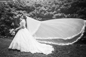 A flowing wedding veil catches the wind in a dramatic black and white outdoor portrait at a garden setting.