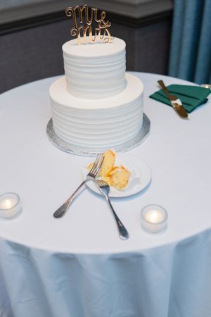 Two-tiered white wedding cake with textured frosting and gold monogram topper on round table with tealight candles.