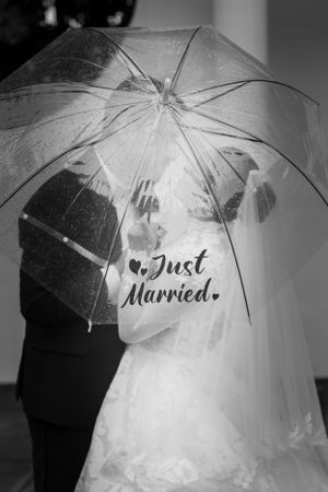 A clear wedding umbrella with 'Just Married' text shelters a couple under a veil in black and white photography.