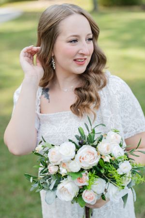 Beautiful bridal portrait outdoors with soft waves and a lush bouquet of blush roses and greenery in white lace dress.
