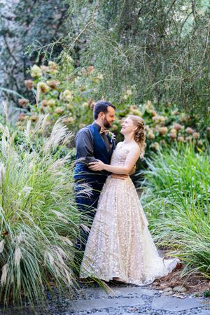 A couple embraces in a romantic garden setting among ornamental grasses and greenery, with bride wearing a lace gown.