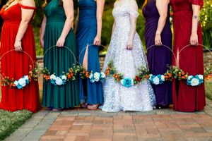 Colorful bridesmaids dresses in red, green, blue, white, and burgundy shades with matching floral bouquets on brick ground.