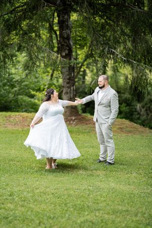 A couple in wedding attire dance together on a lush green lawn surrounded by trees.