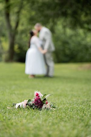A bouquet of red and pink flowers lies on green grass while a couple sits blurred in the background.