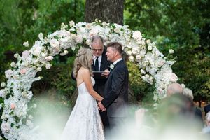 A romantic outdoor wedding ceremony under a floral arch with white roses and greenery in a garden setting.