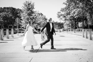 Newlyweds share a playful moment walking together on a tree-lined pathway in black and white.