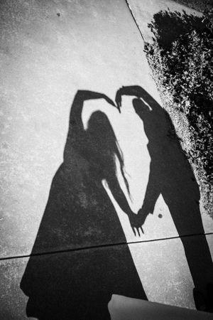 Two shadows cast on ground forming a heart shape with their arms in a black and white artistic silhouette.