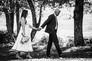 A black and white wedding photograph shows a couple walking hand in hand along a tree-lined path outdoors.
