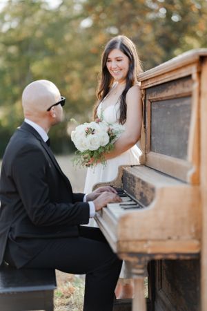 A musician plays a rustic upright piano outdoors while someone in a white dress holds a bouquet of white flowers.