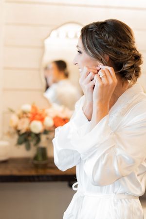A bride getting ready on her wedding day puts on earrings while wearing a white silk robe near floral arrangements.