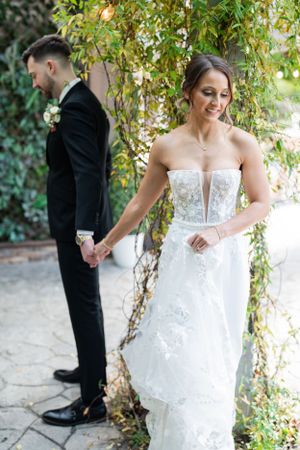 A bride and groom hold hands while standing apart in a garden setting with climbing vines and stone path.