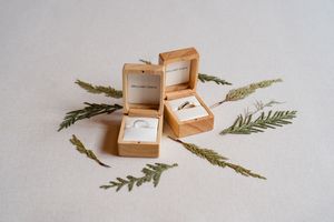 Small wooden ring box with delicate green foliage arranged around it on a light neutral surface.