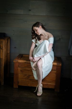 A figure in a white dress sits contemplatively on a wooden storage chest in a dimly lit room.