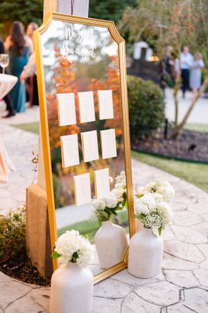 Gold framed mirror seating chart display with white ceramic vases and flowers at an outdoor wedding reception.