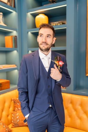 Groom in navy blue three-piece suit with orange and burgundy boutonniere poses against blue built-in shelves and orange couch.