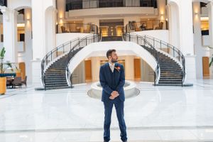 Someone in a navy blue suit stands in an elegant lobby with dual curved staircases and white marble floors.