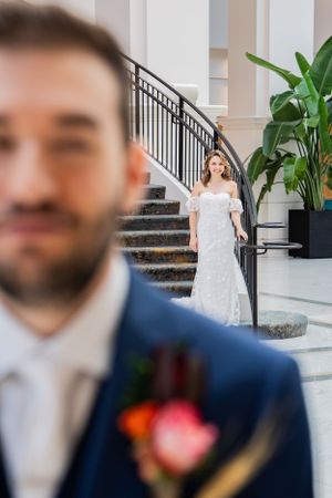 Bride in white lace gown descends a grand staircase while groom in navy suit stands in foreground of elegant venue.