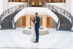 Two people in formal attire dance and embrace in front of an elegant double staircase with an arched design.