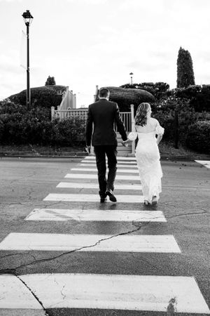 A couple in wedding attire walk hand in hand across a zebra crossing in a black and white photograph.