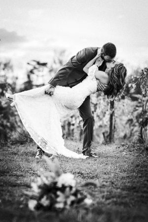 Black and white wedding photo showing romantic dip kiss with bride in lace dress and groom in dark suit in garden setting.