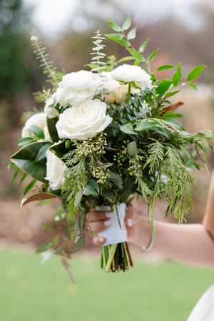 A beautiful bridal bouquet featuring white roses and mixed greenery including eucalyptus and evergreen stems tied with ribbon.