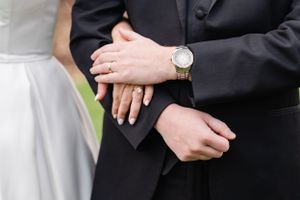 A close-up of newlyweds' hands showing wedding rings and a gold watch against black suit and white dress.