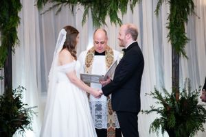 Wedding ceremony under evergreen garland archway with bride in white dress, groom, and officiant during vows.