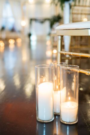 Glowing white pillar candles in tall glass cylinders create romantic ambiance on a dark surface with bokeh lights in background.