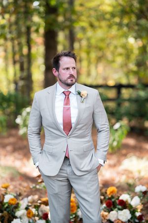 A groom in a light gray suit and red tie poses in a garden with orange and white flowers.