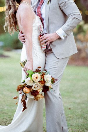 Close up of wedding couple embracing with autumn-colored bouquet featuring roses and dried flowers.