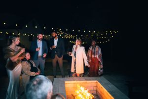 A group of people gather around a bonfire at night with string lights visible in the background.