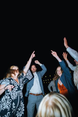 Wedding guests dancing and celebrating with raised arms against a dark background at a reception party.