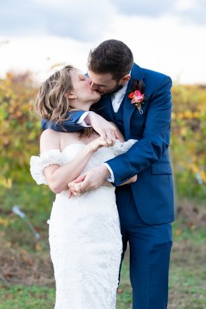 Couple in wedding attire sharing romantic kiss outdoors, with groom in blue suit and pink boutonniere.