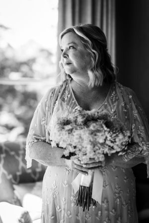 Black and white portrait of a bride in a delicate lace dress holding a bouquet while gazing upward near a window.