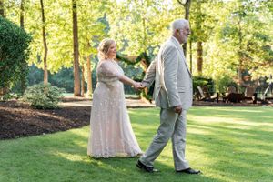 An elder couple wearing gray and blush wedding attire walk together in a sunlit garden setting during their summer wedding celebration.