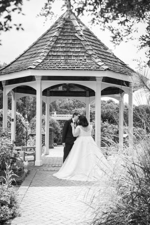 A romantic moment captured in black and white under an elegant wooden gazebo surrounded by lush garden landscaping.