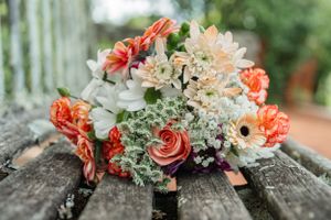A delicate bridal bouquet featuring coral roses, peach gerbera daisies, white baby's breath, and cream blooms on rustic wood.