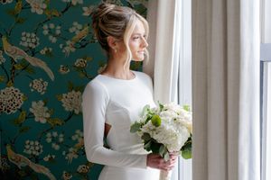 Woman in long sleeve white dress holds white floral bouquet against teal floral wallpaper in sunlit room.