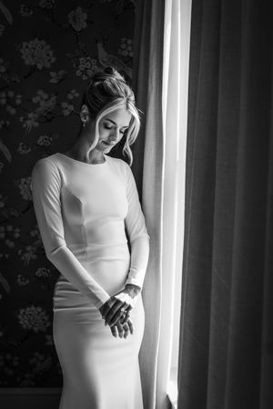 A person in a white long-sleeved dress poses by a window with floral wallpaper in a black and white photograph.
