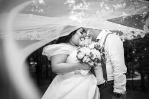 A romantic black and white wedding photograph showing a tender moment beneath a flowing veil.