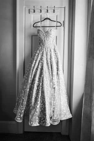 A strapless white lace wedding dress with a full skirt hangs elegantly on a hanger against a white wall.