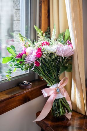 A beautiful bouquet of pink and white flowers tied with a pink satin ribbon sits on a wooden windowsill.
