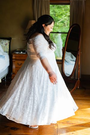 A white lace wedding dress with a full skirt twirls gracefully on a hardwood floor in natural window light.