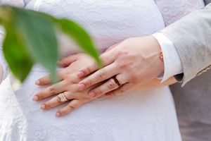 Close-up of hands with wedding rings holding green leaves against a light background.