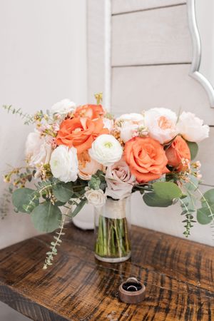 Beautiful floral arrangement with coral roses, white ranunculus and eucalyptus in a glass vase on rustic wooden table.
