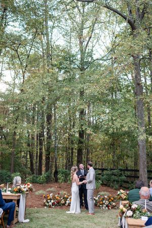 A romantic outdoor wedding ceremony takes place in a wooded setting with autumn floral arrangements.