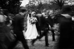 Black and white artistic wedding photo showing figures in motion blur moving around a bride holding a bouquet.